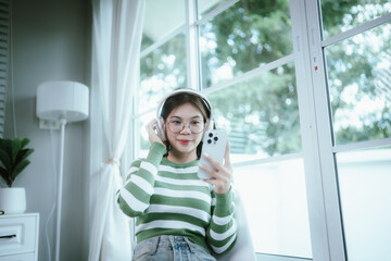A young woman in a striped shirt and glasses relaxes indoors, enjoying music or a podcast through...