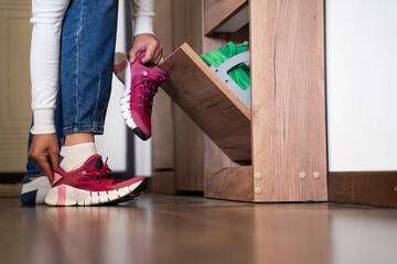 Unrecognizable woman putting on pink sneakers on her feet in hallway indoors, home interior background, daily routine lifestyle, wear casual outfit, modern interior, storage and tidiness concept