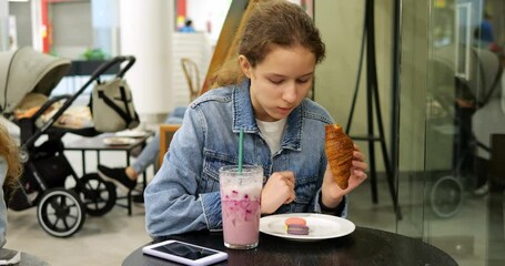 Young girl bites croissant enjoying snack moment at modern cafe. Fresh pastry pairs with vibrant drink standing beside phone and macaron - Powered by Adobe