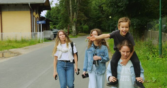Family group walks along road in natural setting with boy happily riding on sister shoulders with keychain. Mum and daughter suggest leisurely outdoor journey