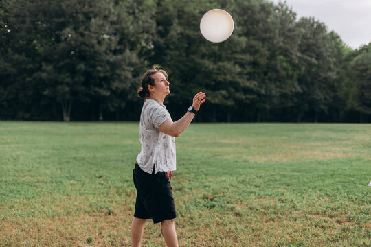 Man in profile view with hair tied back about to catch or throw frisbee disc hovering air open green space trees in soft blurred background relaxed active summer day