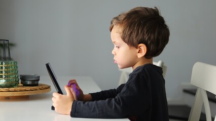 Smart preschooler boy touches screen of tablet with finger to open app sitting at desk. Little kid...