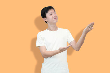 A young Asian man in a white t-shirt points upwards with palms against an peach background,...