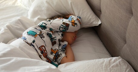Little boy in pajamas with cars lies in bed with soft linen covering head with hand. Carefree child...