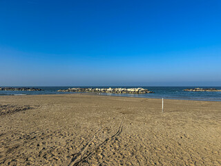 The wide, empty sandy beach of Misano Adriatico in winter, with tire tracks on the sand and distant buildings under a clear blue sky.