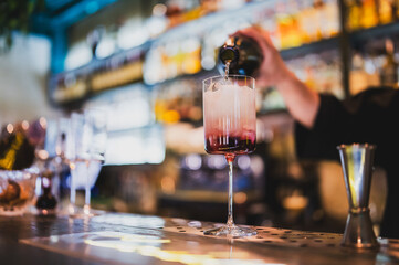 Bartender pouring a vibrant cocktail into a tall glass with ice at a bar. Colorful drink with gradient tones, professional bar service, blurred alcohol bottles in background.