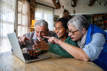 Joyful multiethnic grandparents with grandchild watching online game on laptop at home together