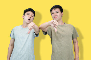 Two young Asian men stand against a yellow background, giving thumbs-down gestures with serious...