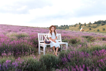 Beautiful small girl in white dress collects lavender on lavender field. Cute child girl in straw boater hat sit on bench in the middle of lavender bushes with basket violet flowers. Provence, France.