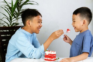 Two happy Asian boys laughing as they feed each other delicious red velvet cake with spoons in a...