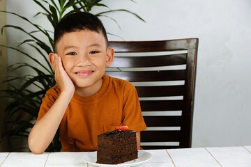 Cute boy in orange shirt smiling while sitting at a table with a slice of chocolate cake in front of him. Joyful childhood moment with dessert at home.
