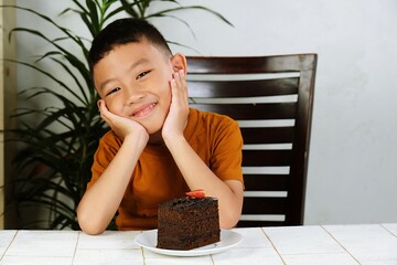 Happy young boy smiling sweetly with both hands on cheeks, sitting at a table with a slice of chocolate cake. Adorable indoor moment of childhood joy and dessert.