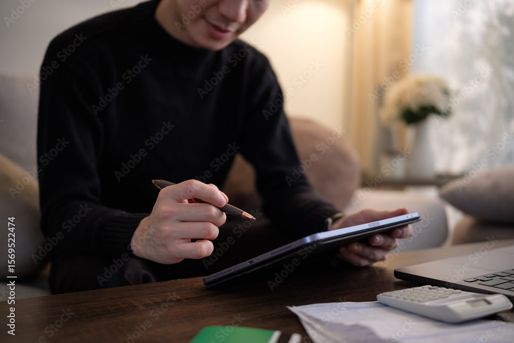Poster man working on digital tablet with stylus while reviewing financial documents at home.