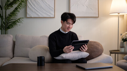 Young man sitting on a cozy couch and using a digital tablet at home. Relaxed and focused in a modern living room environment.