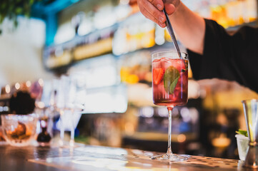 Bartender stirring a refreshing cocktail with strawberries, mint, and ice in a tall glass on a bar...