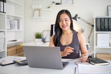 Smiling young woman working on a laptop at her desk, surrounded by charts, reports, and office...