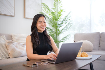 Fototapeta premium Smiling young woman working on a laptop at home with breakfast on the table.