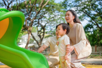 Caring parents encouraging their toddler near a playground slide during a sunny day at the park.
