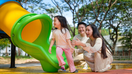 Happy Asian family enjoying playtime with their daughter at a colorful playground slide.