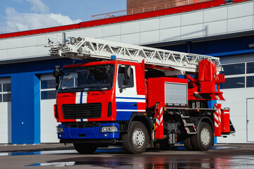 A vibrant red and white fire engine with an extended ladder is parked on a paved surface in front of a fire station.
