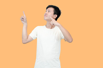 A young Asian man in a white t-shirt points upwards with both index fingers against an peach...