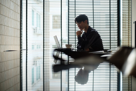 Young man working on a laptop in a stylish office with window blinds and reflections.