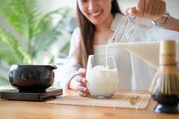 Smiling woman pouring milk into a glass for making iced matcha latte at home.