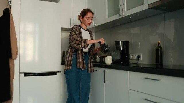 A woman pours coffee from a coffee pot into a cup and drinks while looking at her phone