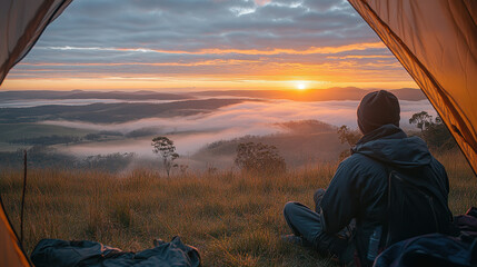 A man sits in a tent looking out at a beautiful sunset. The sky is filled with clouds and the sun is setting in the distance. The man is wearing a black jacket and a hat