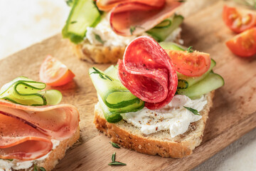 Delicious sandwiches with cream cheese, salami, cucumber, tomatoes on wooden board close up on light background. Homemade appetizer
