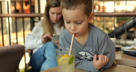 Obraz premium Son drinks lemonade with straw savoring flavor against mom using phone. Family shares quiet moment in restaurant with mother attention fixed on device