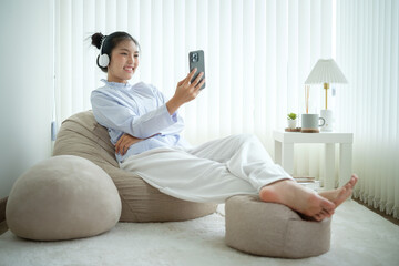 Relaxed Asian woman listening to music and using mobile phone while sitting comfortably by a large...