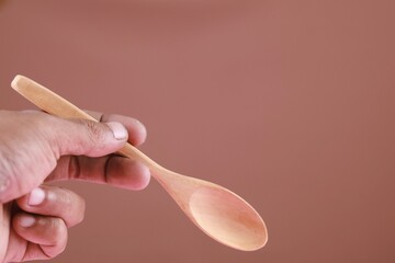 Close up hand holding wooden spoon, eco friendly cutlery isolated on brown background. Conscious...