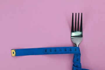 Stainless fork tied with twisted blue measuring tape isolate on pink background. Diet and fit body...