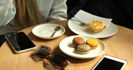 Woman sits at cafe table with cup of coffee macarons and slice of pastry. Coffee break and sweet indulgence with smartphones and sunglasses