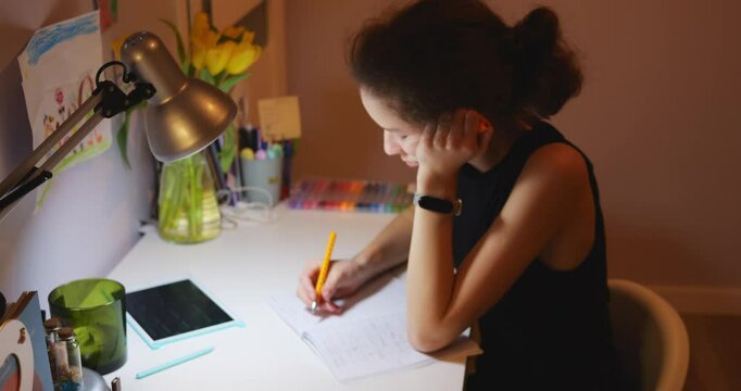 Senior schoolgirl studies hard at home and does homework in evening. Student writes in paper notebook and focuses on task under bright light of desk lamp