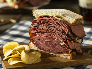 Thick pastrami sandwich stacked with beef slices and cheese on rye bread, served with potato chips on a wooden board, set on a checkered cloth in warm lighting
