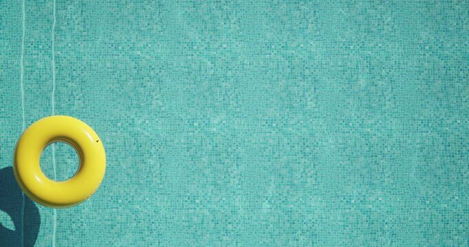 Top view of a bright yellow inflatable ring floating on clear turquoise water in a swimming pool. Sunlight casts a distinct shadow on the mosaic tiles. Minimal, peaceful summer vacation vibe