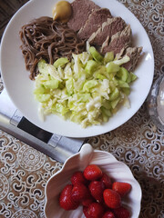 A plate of sliced meat, noodles, and chopped vegetables. A bowl of fresh strawberries is nearby....