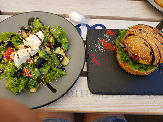 A fresh salad with feta cheese and a gourmet burger on a black slate plate, includes greens,...