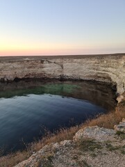 A serene natural pool surrounded by rocky cliffs at sunset. The calm water reflects the colors of the sky, creating a tranquil atmosphere.