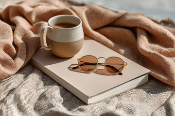 Cozy scene featuring a warm cup of coffee, stylish sunglasses, and a soft blanket draped over a neutral book