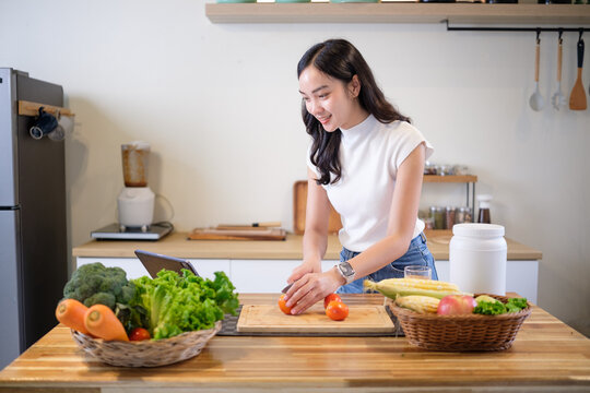 Asian woman standing at kitchen counter slicing tomatoes with fresh produce around her. Concept of clean eating, healthy lifestyle, and meal preparation.