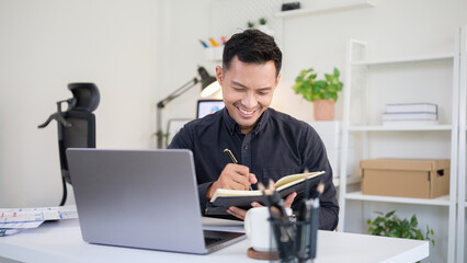 Happy businessman taking notes in a notebook while working with laptop and paperwork in an office.