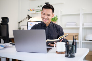 Happy businessman taking notes in a notebook while working with laptop and paperwork in a office.