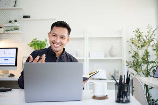 Smiling businessman holding a notebook while having a video call meeting on his laptop in a modern office.
