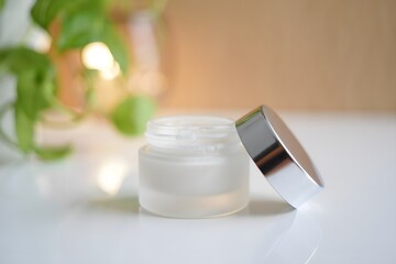 Frosted glass jar of cream with silver lid and blurred green plant background