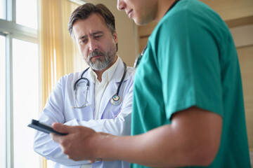 Senior male doctor gives clear advice to a Male nurse while checking patient data on a tablet in...