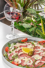 A gourmet beef carpaccio served with parmesan shavings, arugula, capers, cherry tomatoes, and lemon, paired with a glass of red wine in an elegant setting