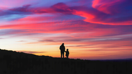 Fototapeta premium A dramatic landscape photograph of a silhouetted adult and child standing on a dark hillside against a spectacular sunset sky.